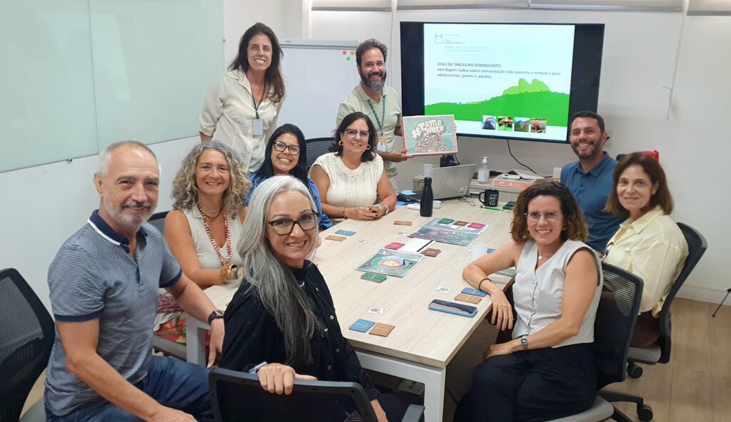 Foto dos profissionais do encontro na sala da Escola Corporativa Fiocruz sentados em volta de uma mesa em que está o tabuleiro e cartas do jogo tamojunto