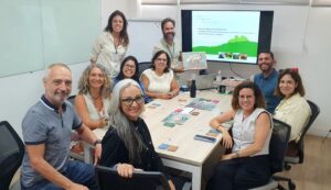 Foto dos profissionais do encontro na sala da Escola Corporativa Fiocruz sentados em volta de uma mesa em que está o tabuleiro e cartas do jogo tamojunto